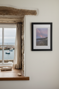 Rainbow Over The Ship coastal photography print in a black frame, displayed on a whitewashed stone wall in a charming fisherman's cottage with a sea view through the window